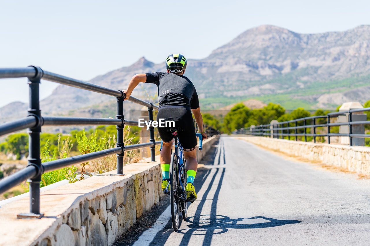 Back view of unrecognizable bicyclist in sportswear and helmet resting near railing while riding bike up paved road in rural mountainous area