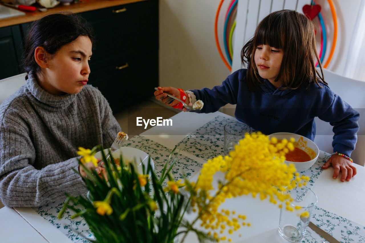 Two sisters eating lunch at home at white table with yellow flowers