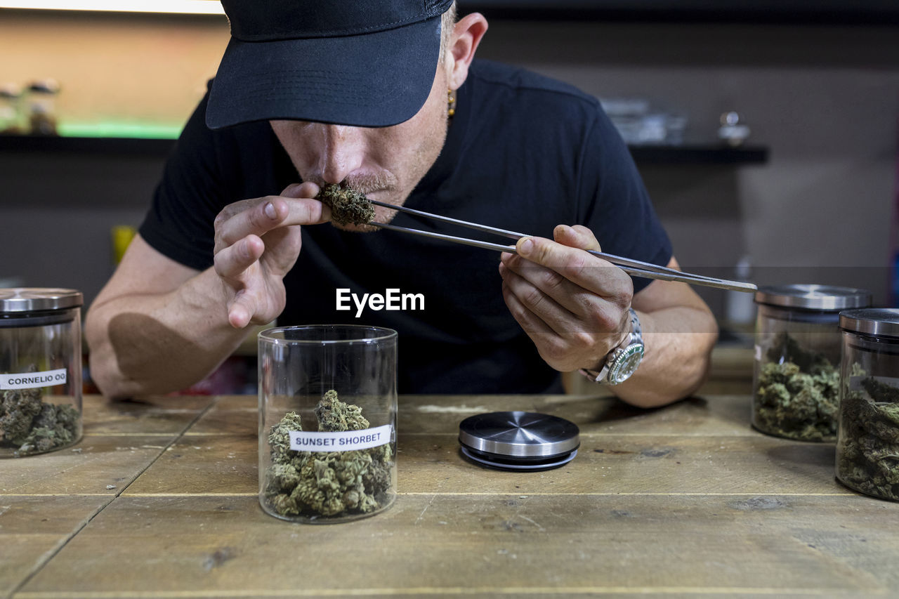 Unrecognizable adult male in cap with tweezers smelling dried hemp floral bud above table with jars in workspace
