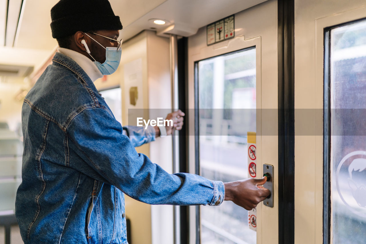 Side view of african american male passenger in mask standing at doors and pressing button before leaving train while traveling during coronavirus epidemic