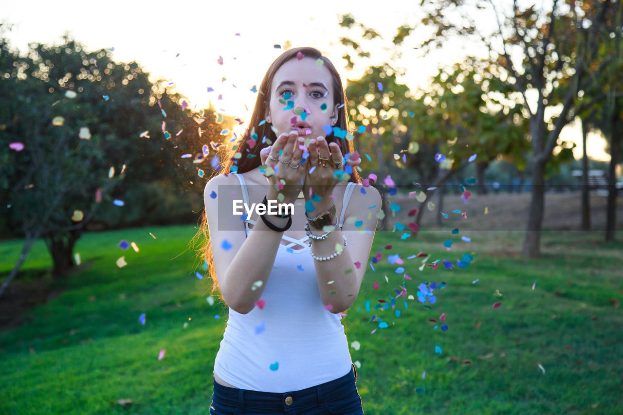 Portrait of young woman blowing confetti while standing at park