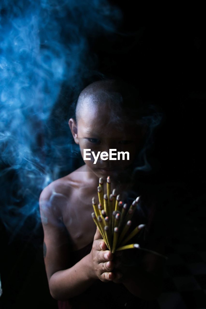 Portrait of shirtless boy holding incense while standing against black background