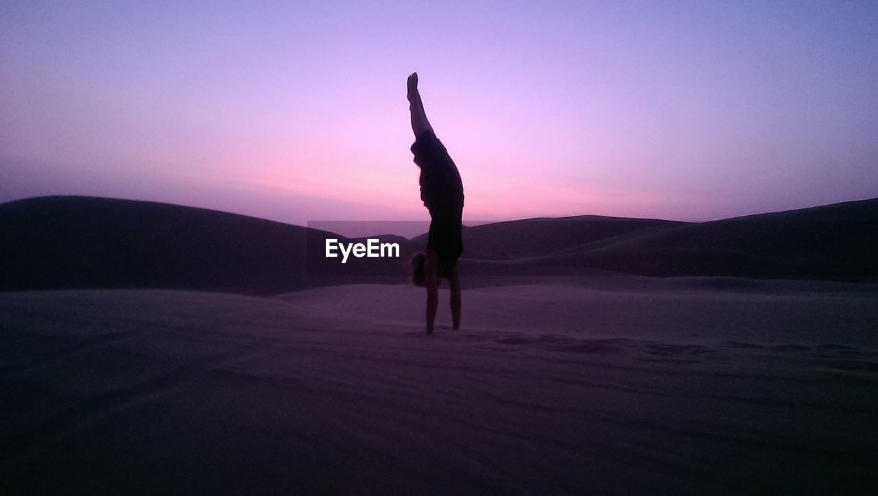 Silhouette man handstand on sand against sky during sunset
