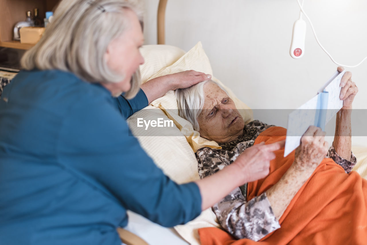 Woman taking care of old woman lying in bed reading book