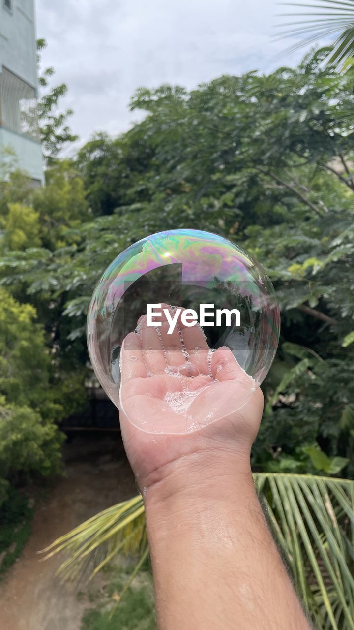 CLOSE-UP OF HAND HOLDING BUBBLE AGAINST BLURRED TREES