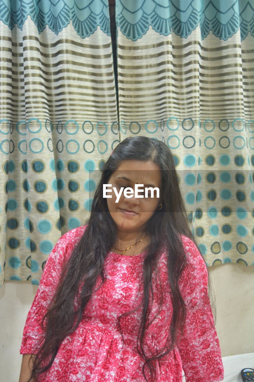 one person, pattern, women, pink, front view, indoors, portrait, smiling, long hair, adult, curtain, standing, hairstyle, textile, waist up, looking at camera, lifestyles, clothing, happiness, emotion, dress, female, young adult, casual clothing, person, red, looking, floral pattern, human face, brown hair, leisure activity