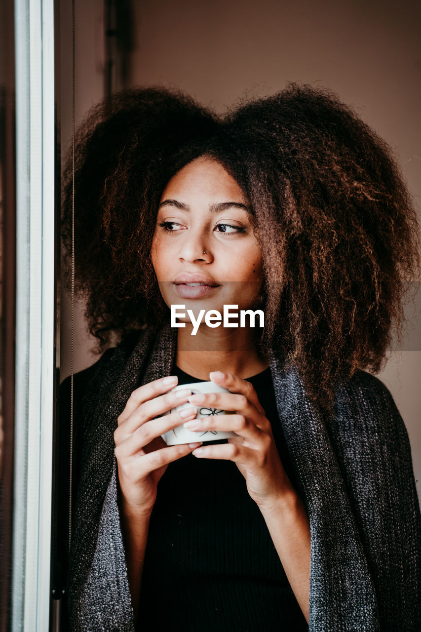 Thoughtful woman drinking coffee while looking through window at home