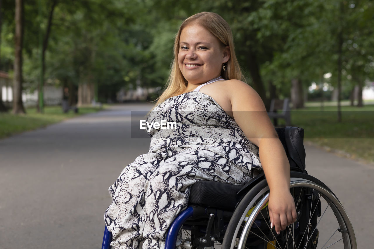 Smiling happy young woman with short stature on wheelchair enjoys time in green park at summer day