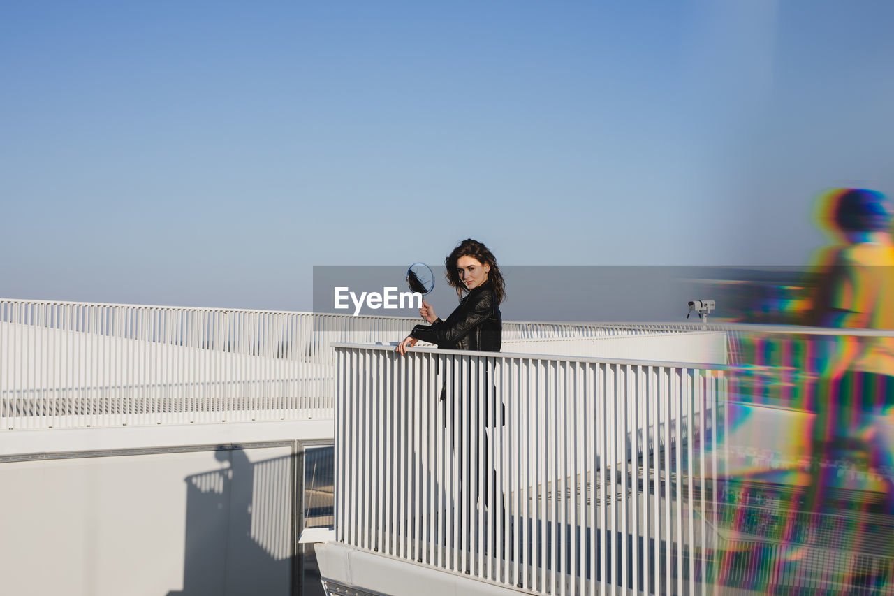 Portrait of woman standing with mirror on building terrace against clear sky