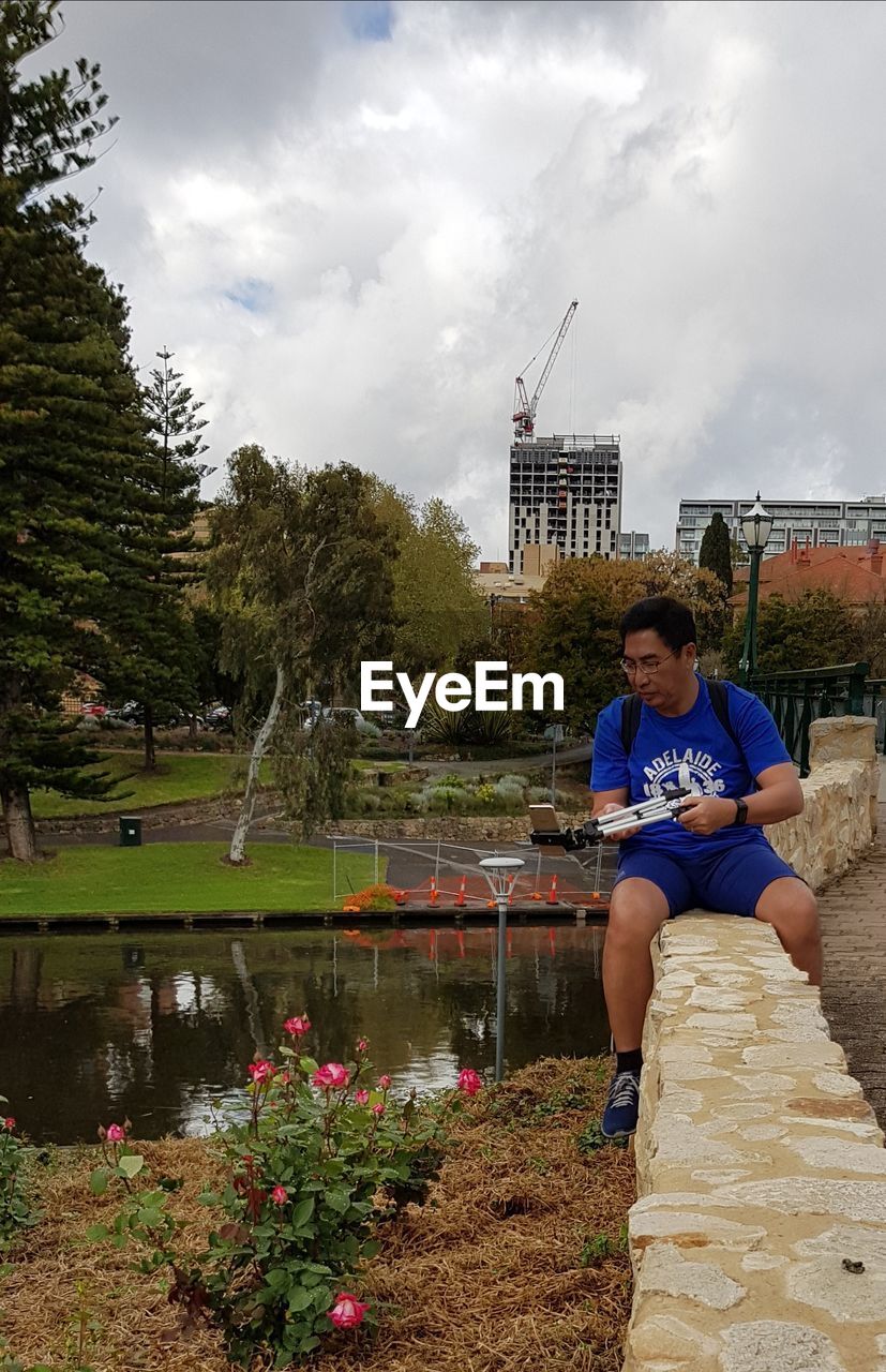 Man with smart phone and tripod sitting on retaining wall in park against sky