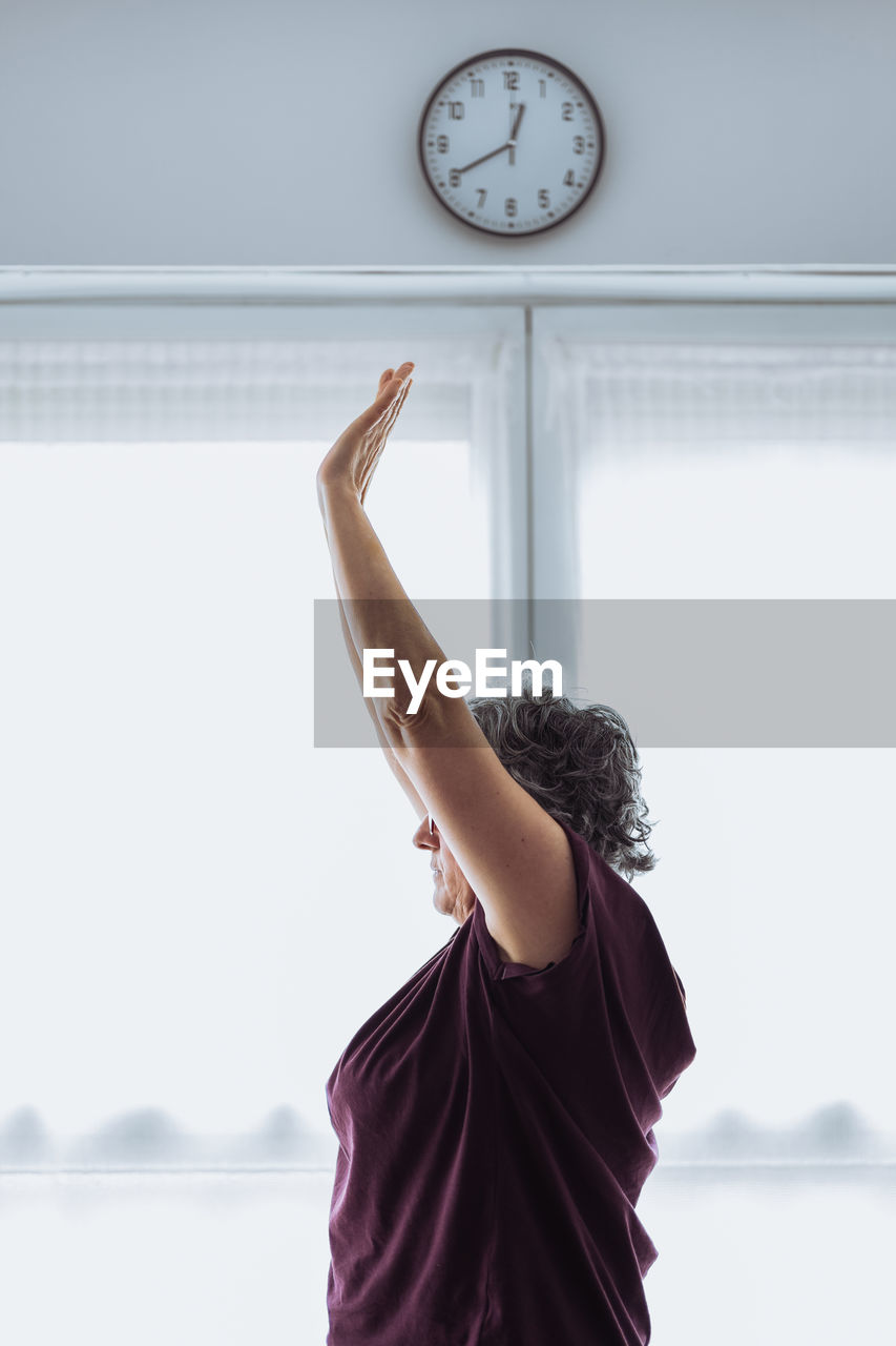 Side view of elderly female in activewear with short gray hair raising arms while doing stretching exercise against window in sunlit room in morning at home