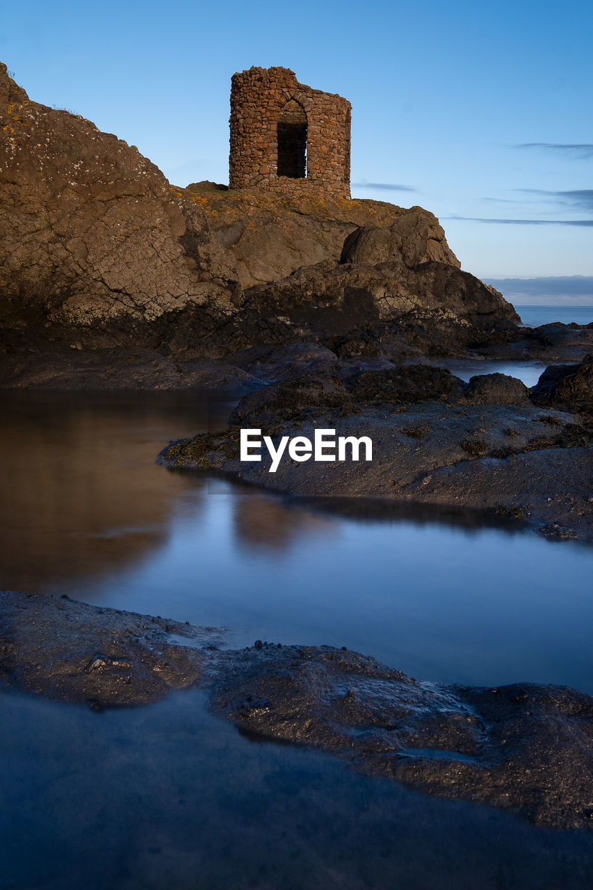 reflection, water, sea, rock, sky, architecture, history, coast, land, travel destinations, nature, the past, landscape, travel, built structure, blue, building, scenics - nature, no people, building exterior, mountain, ocean, environment, beach, outdoors, castle, tourism, tower, ancient, beauty in nature, sunset, evening, clear sky, cloud, dusk