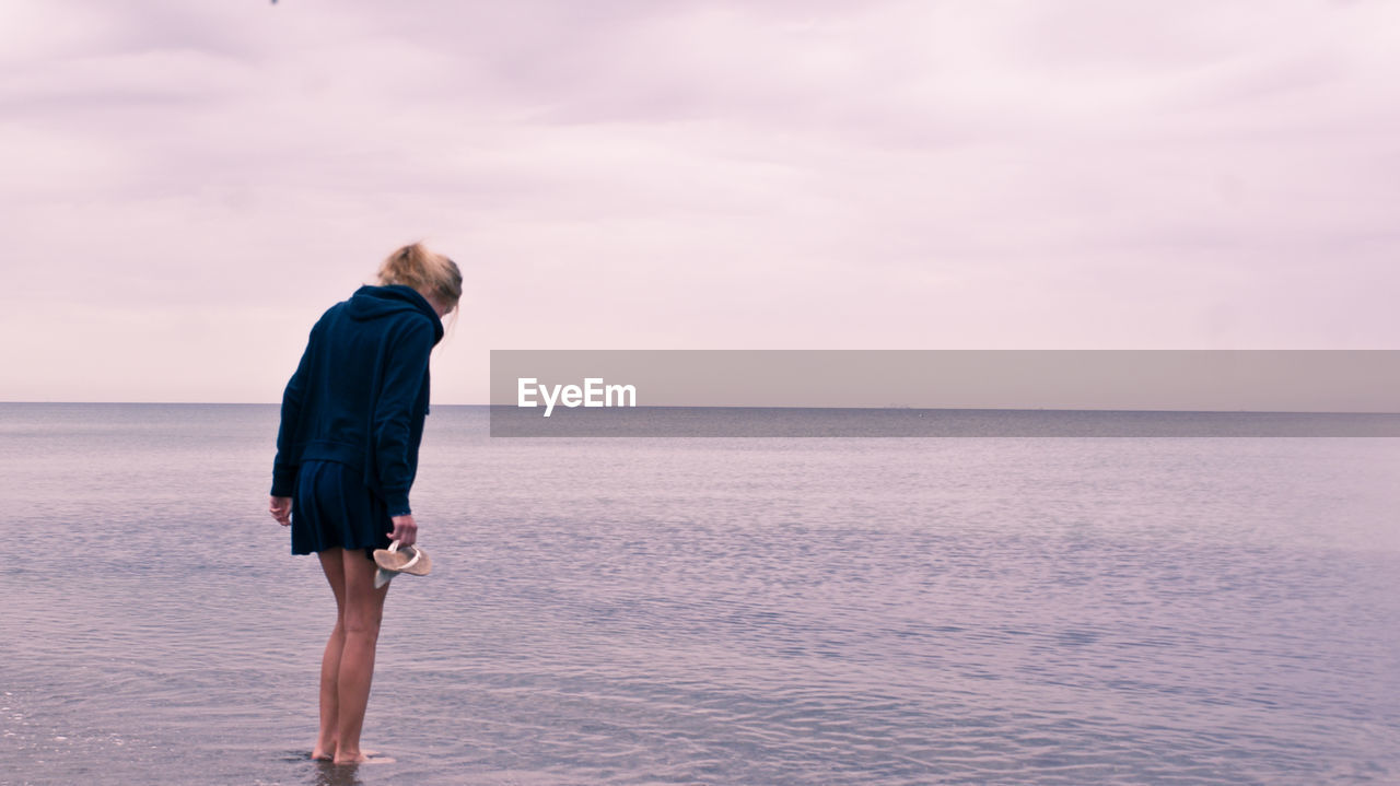 Full length of woman standing in water at beach against cloudy sky