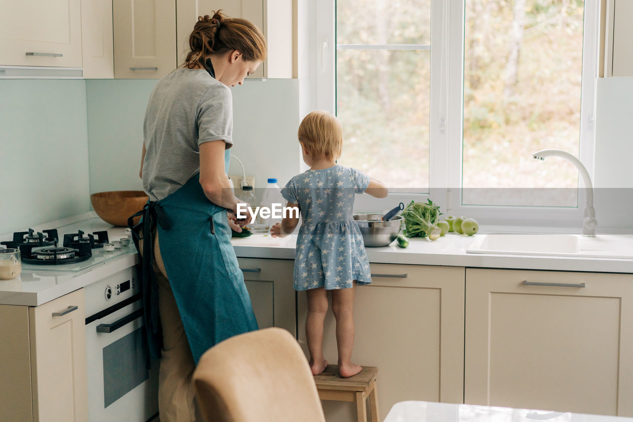 View from the back of a mother with a little daughter cooking in the home kitchen.