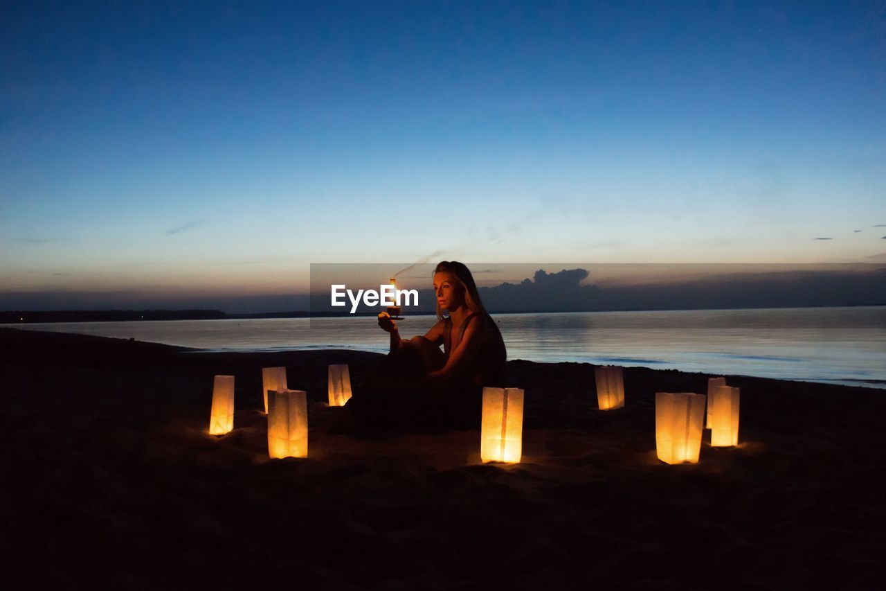 Woman sitting amidst lanterns at beach during dusk