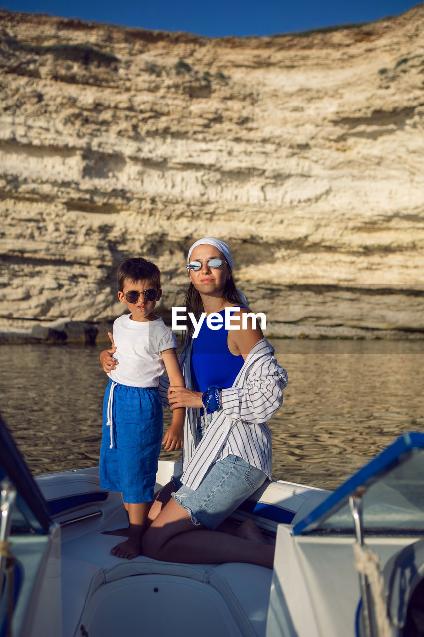 Woman and a boy  in sunglasses are sitting on yacht near rocks in  sea in summer