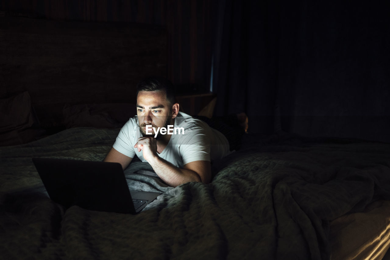 Thoughtful man using laptop while lying on bed at home