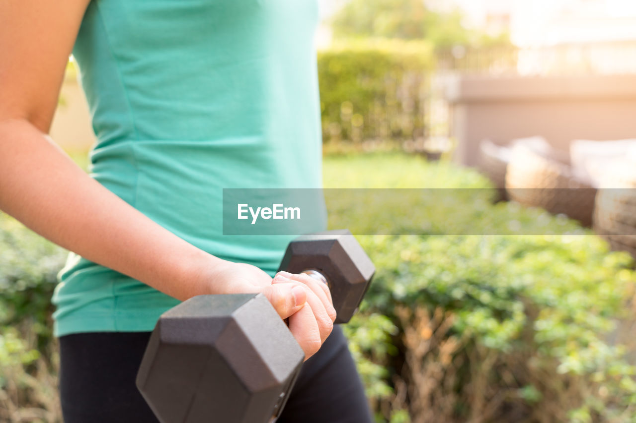 Midsection of woman holding dumbbell while exercising in backyard