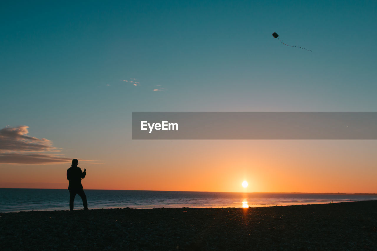 Silhouette man flying kite while standing at beach against sky during sunset