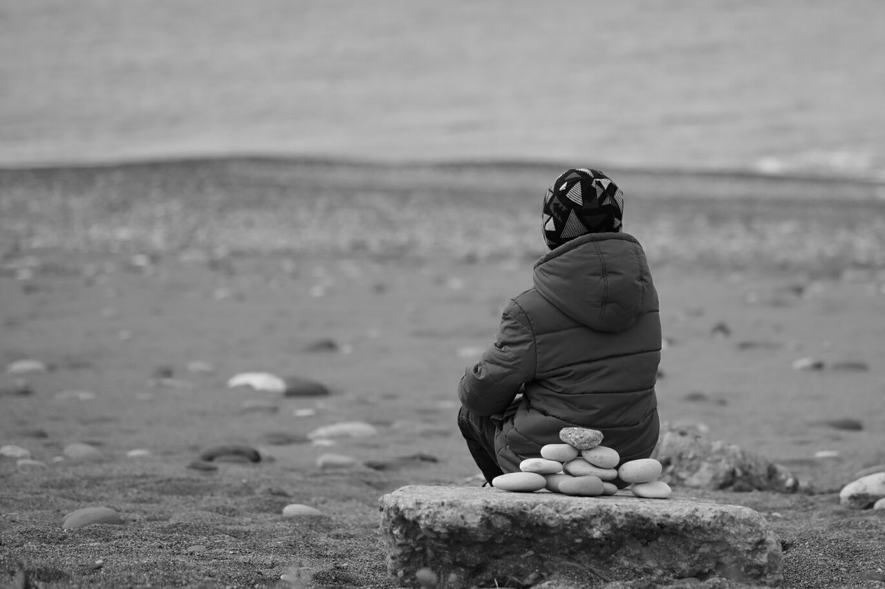 Rear view of hooded person sitting on rock at beach