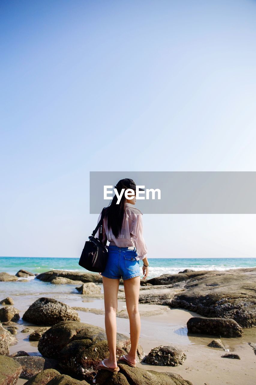 Full length of woman standing on rock at beach against clear sky