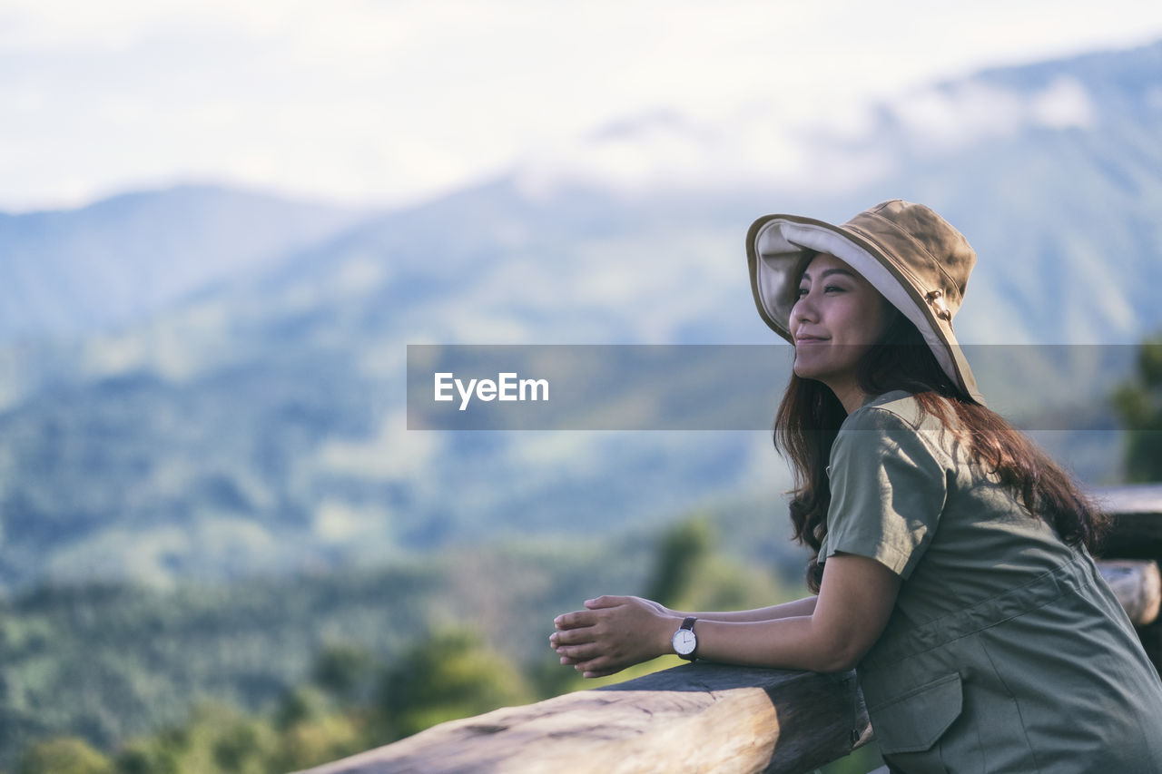 Woman wearing hat while standing by railing against mountain