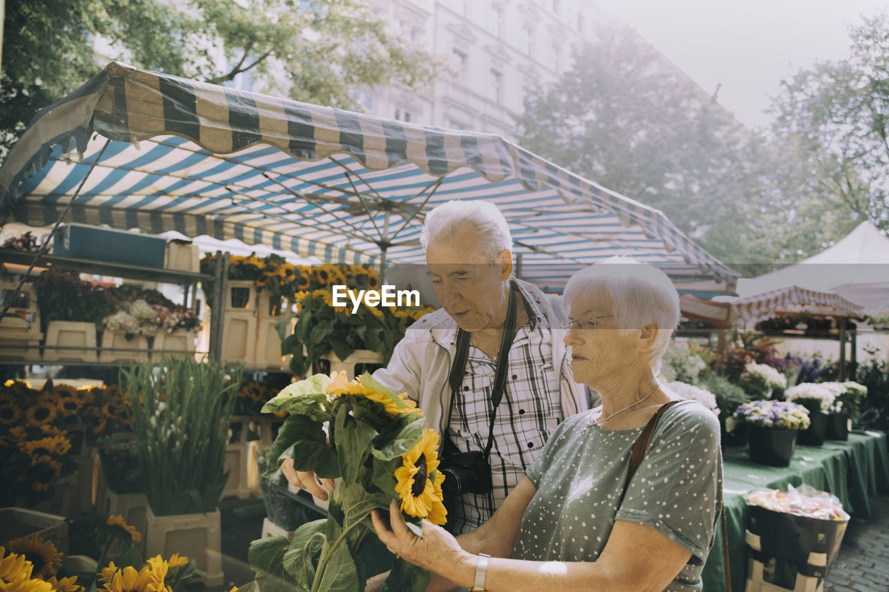 Elderly woman holding sunflowers while shopping with man at flower market in city