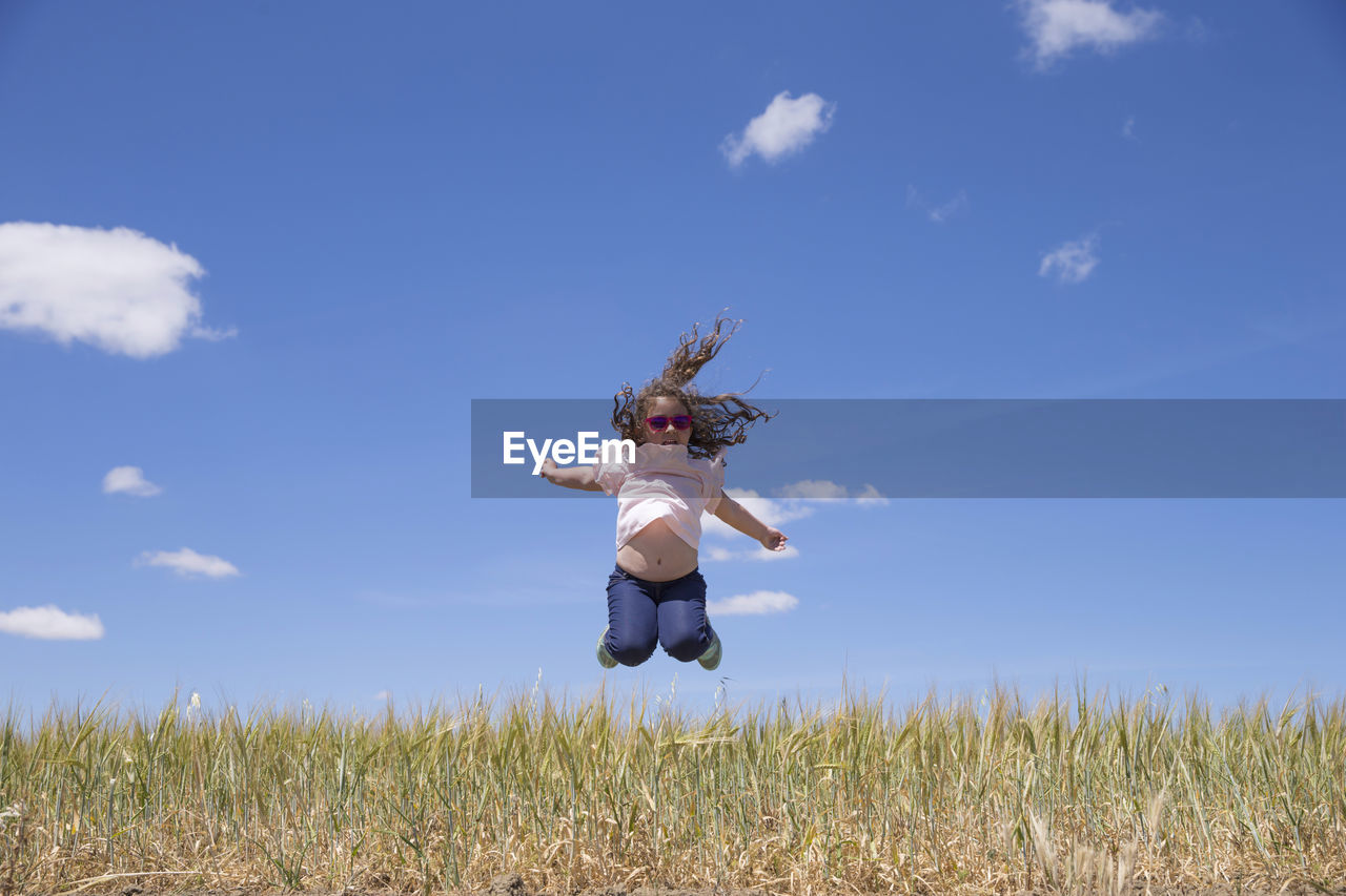 Full length of cheerful girl jumping on grassy field against sky