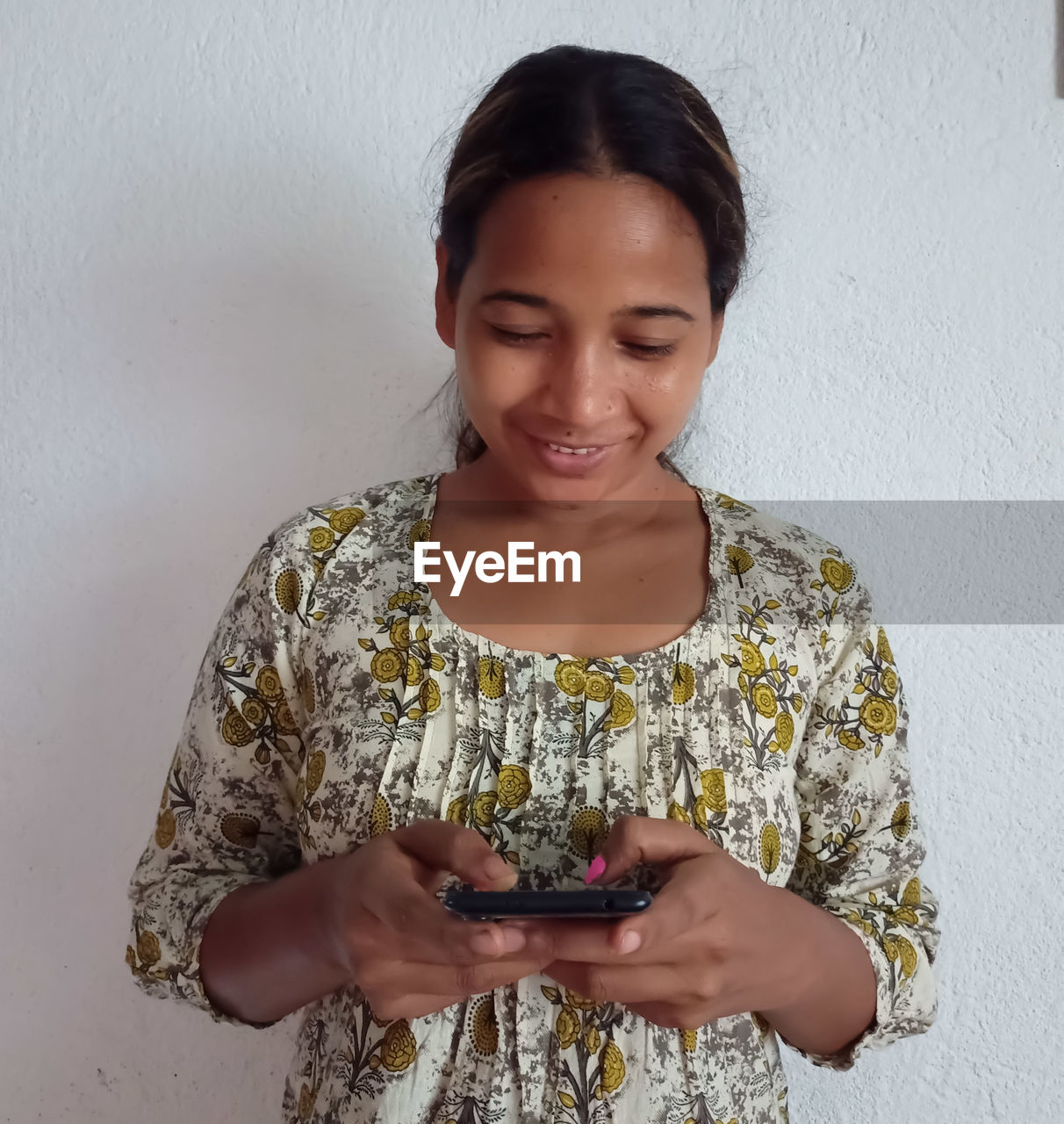 YOUNG WOMAN USING PHONE WHILE STANDING AGAINST WALL