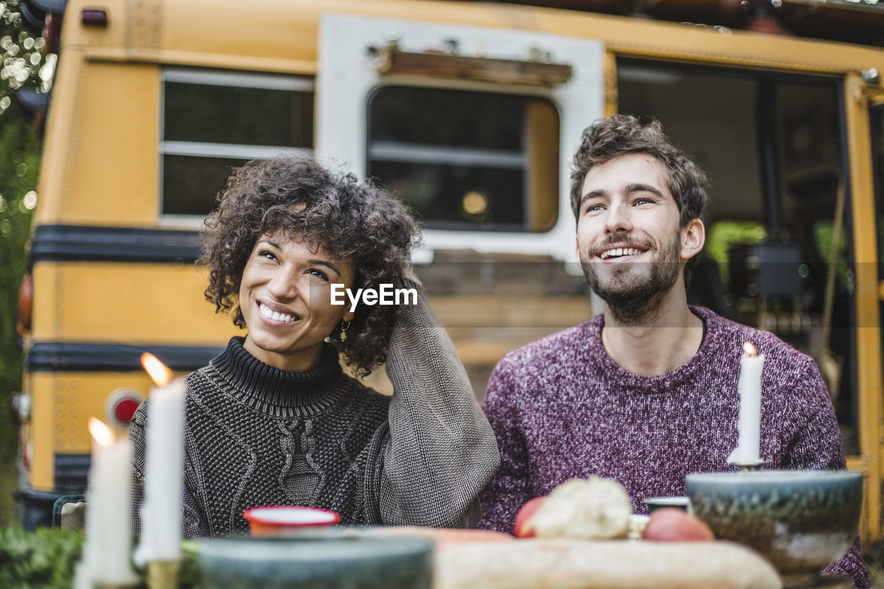 Smiling young couple looking away while sitting at table against caravan during camping