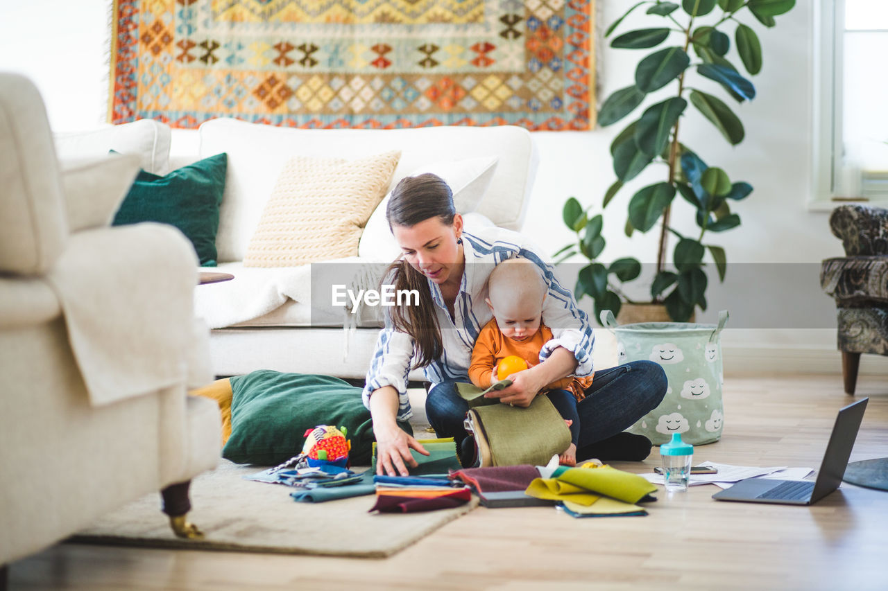 Full length of multi-tasking mother with fabric swatches and daughter working in living room at home