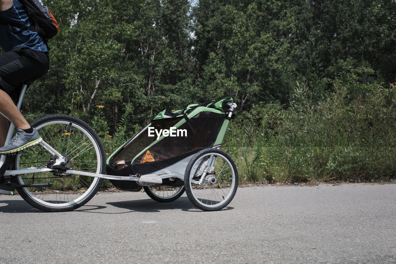 Low section of man cycling with trailer on road against trees during sunny day