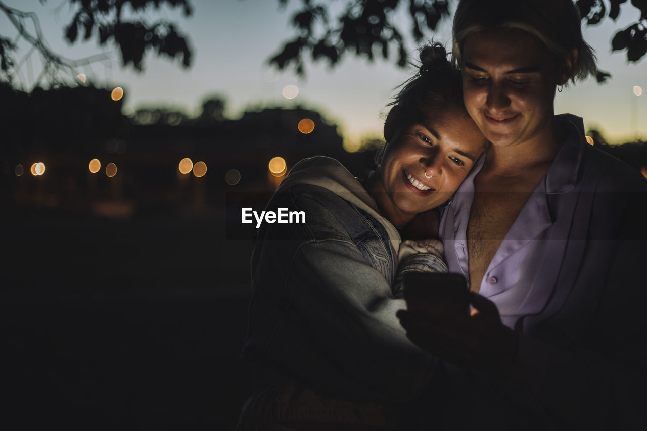 Smiling woman embracing non-binary friend using mobile phone at night