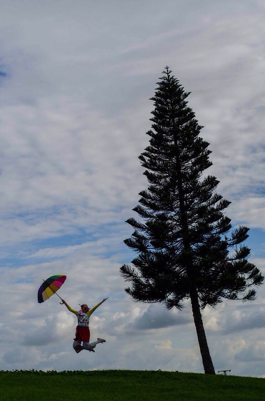 Clown with umbrella jumping on grassy field by tree against sky