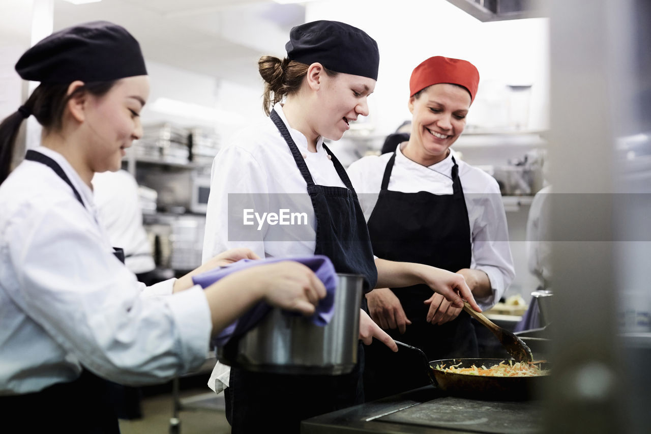 Teacher watching female chef students cooking food in commercial kitchen