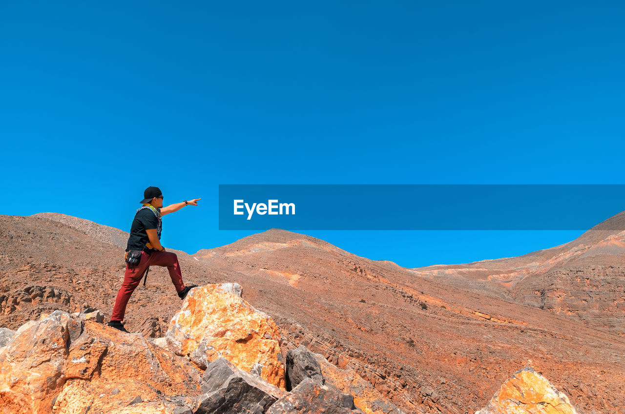 Low angle view of man pointing while standing on rock against blue sky