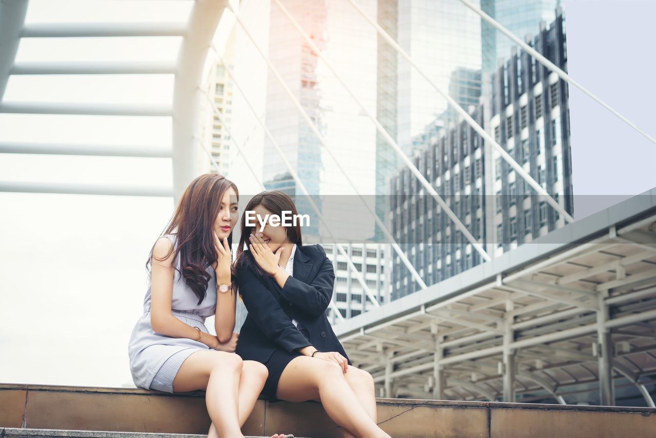 Smiling young woman whispering something with friend while sitting on steps in city