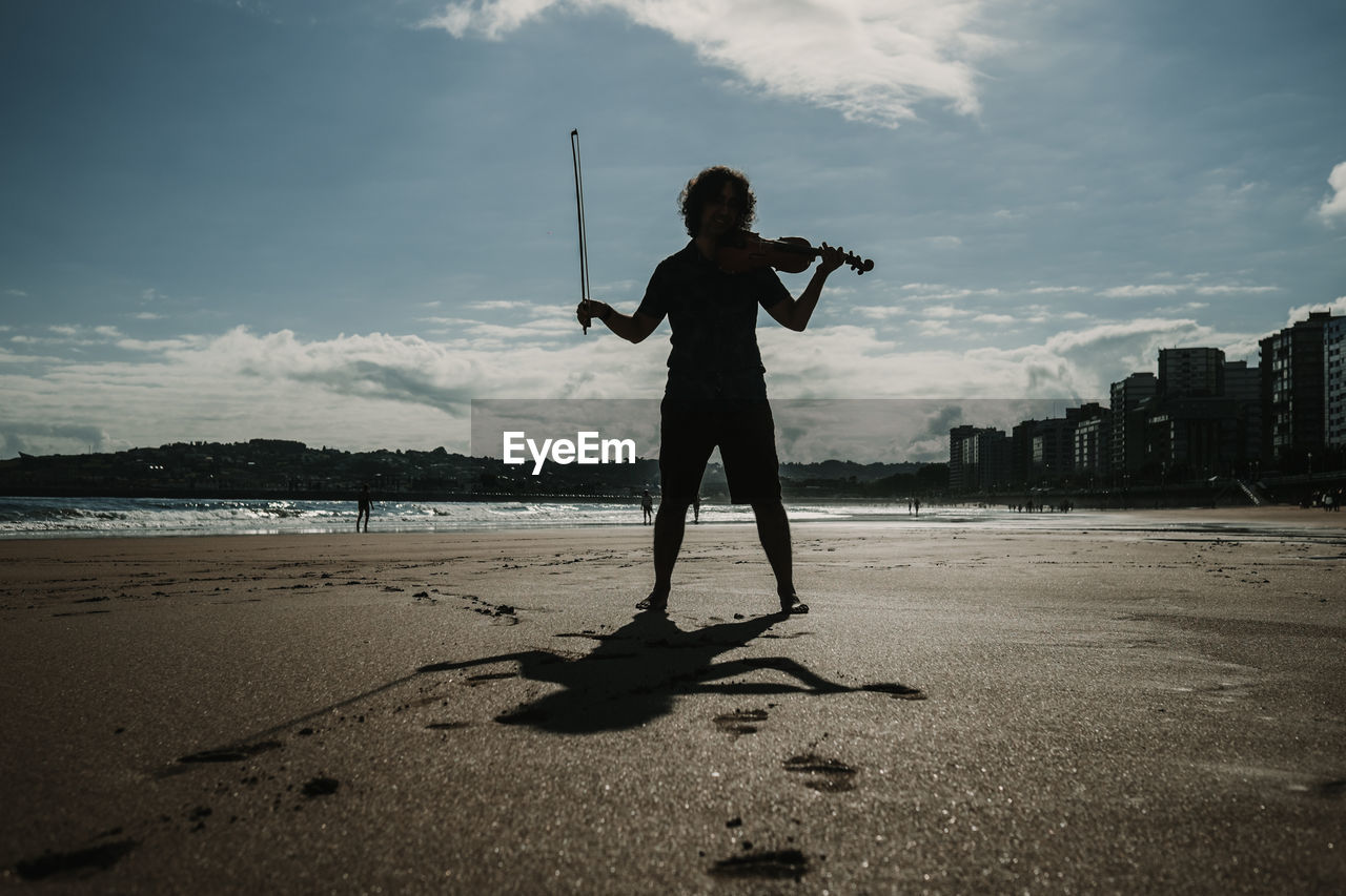 Full length of man playing violin standing on beach against sky