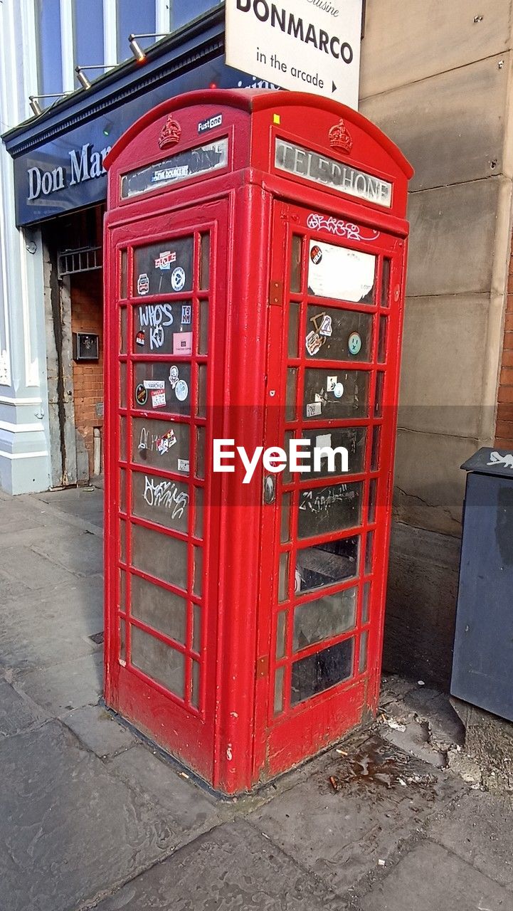 communication, telephone, telephone booth, red, text, architecture, city, western script, street, technology, no people, outdoor structure, convenience, payphone, built structure, building exterior, day, footpath, sidewalk, outdoors