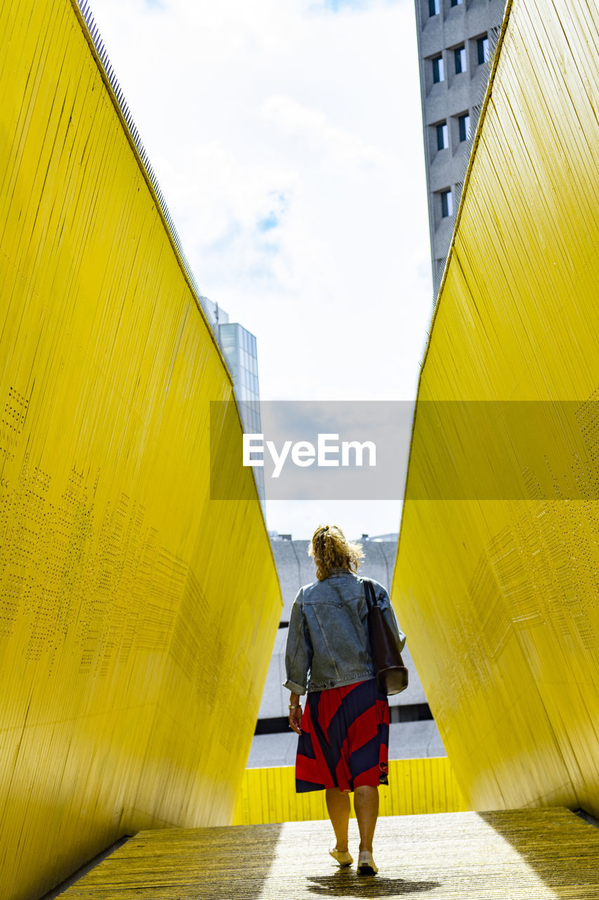 REAR VIEW OF WOMAN STANDING ON UMBRELLA AGAINST YELLOW WALL