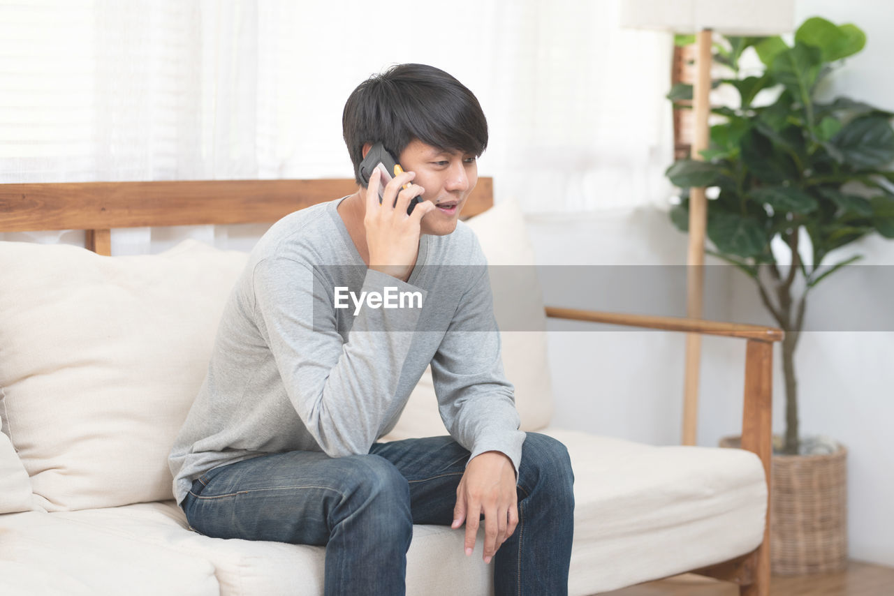 YOUNG MAN USING PHONE WHILE SITTING ON SOFA AT HOME