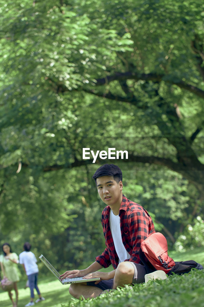 A handsome young asian man sits on the lawn in the summer at the park.
