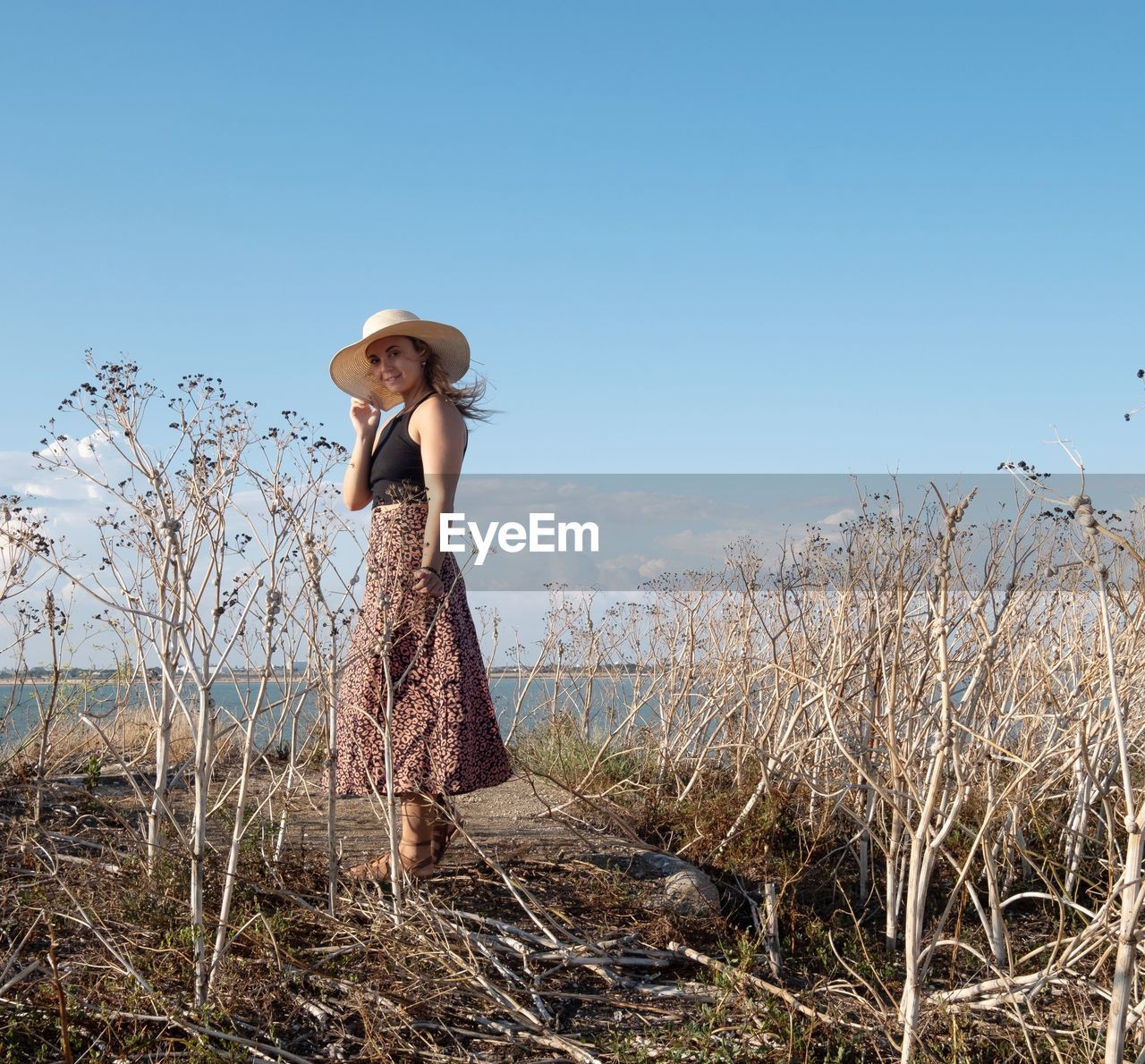 one person, sky, nature, plant, clothing, adult, women, hat, land, landscape, standing, rural scene, grass, agriculture, field, clear sky, full length, day, blue, dress, rural area, environment, young adult, fashion, outdoors, copy space, sunny, tranquility, straw hat, looking, leisure activity, long hair, beauty in nature, sunlight, casual clothing, side view, tree, female, hairstyle, spring, flower, sun hat, lifestyles, farm, summer, solitude, non-urban scene, emotion, fashion accessory, contemplation, scenics - nature