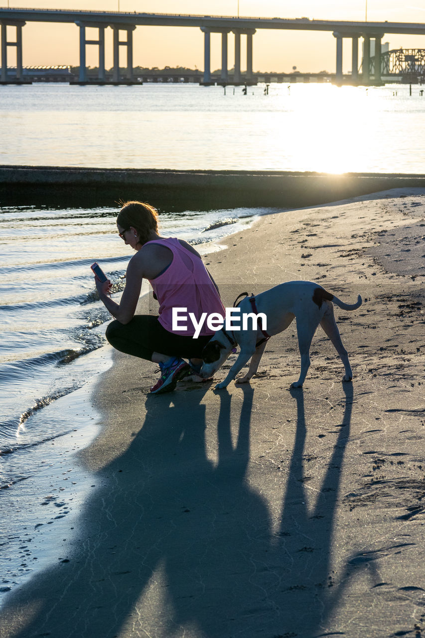 Full length of woman with dog crouching on shore at beach during sunset