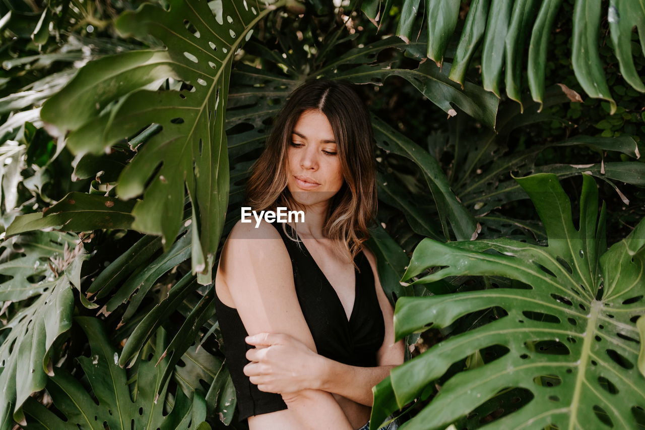 Thoughtful female surrounded by green monstera plants looking at camera while standing in exotic garden in summer