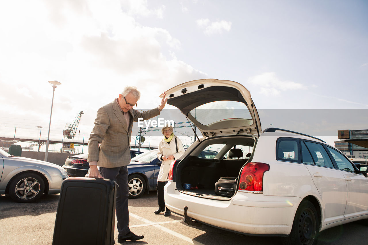 Senior man loading luggage in car trunk with woman standing in parking lot against sky