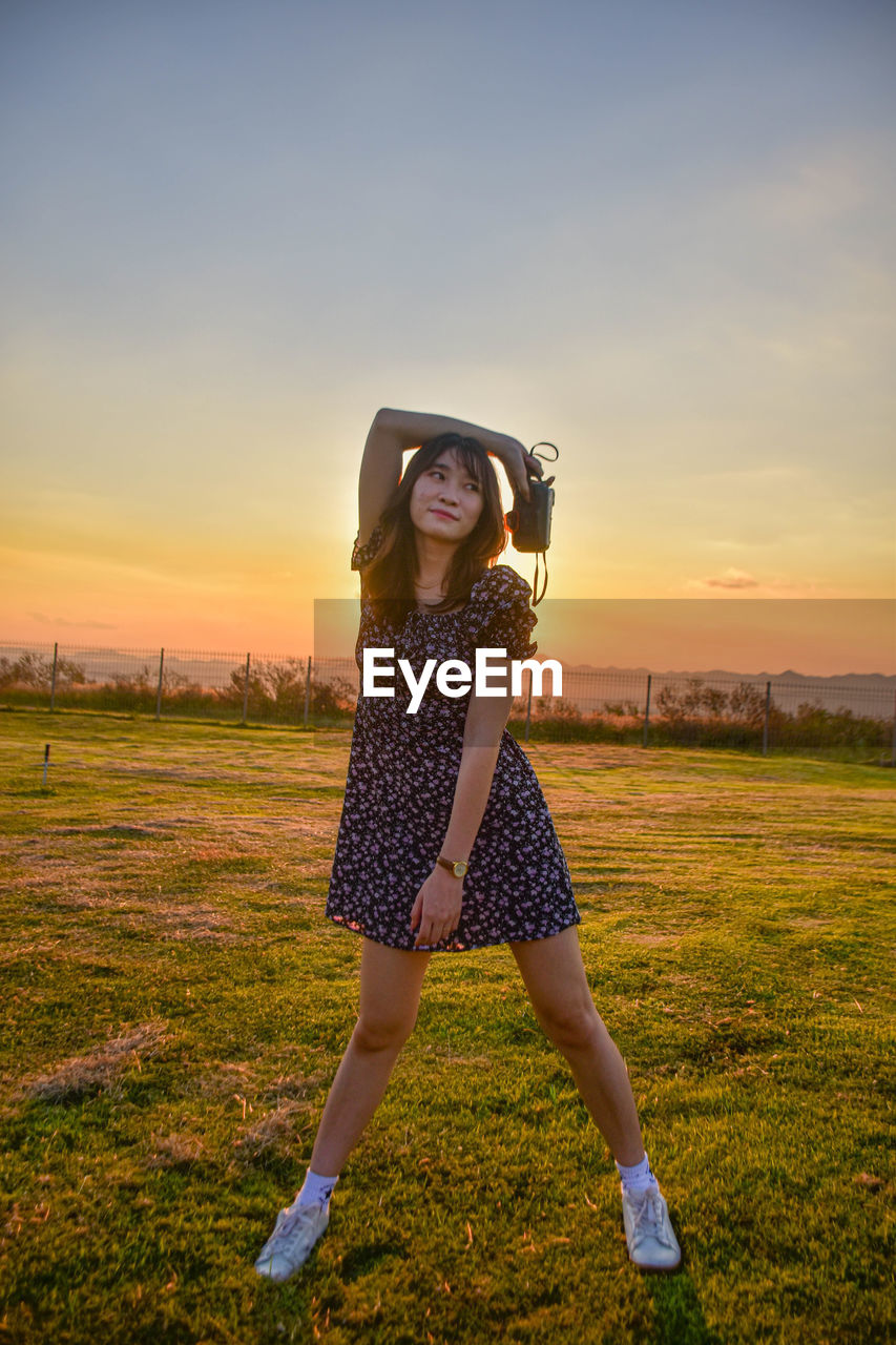 Portrait of young woman standing on field against sky during sunset