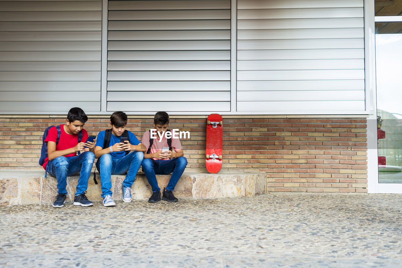 Friends using smart phones while sitting against school building