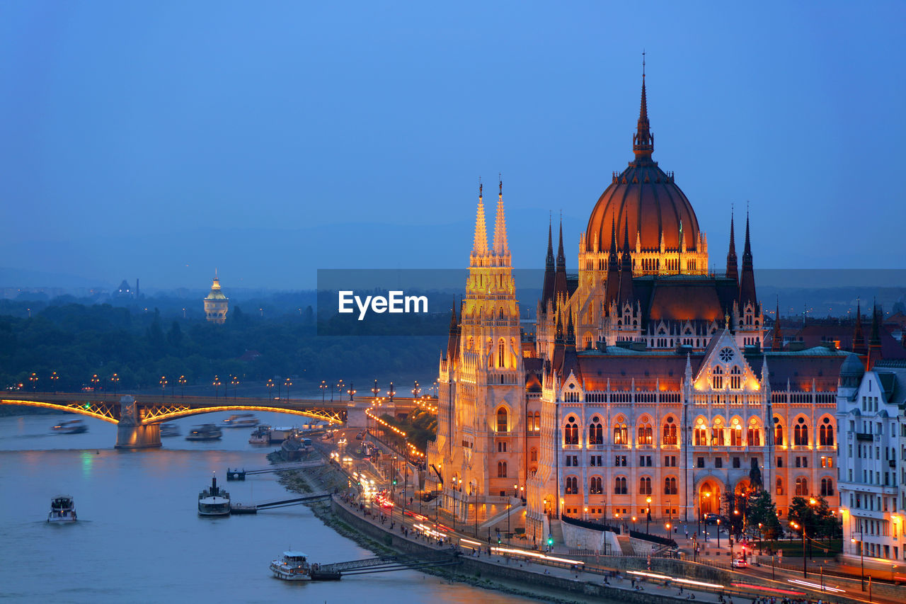 Bridge over river by illuminated hungarian parliament building against sky in city at dusk