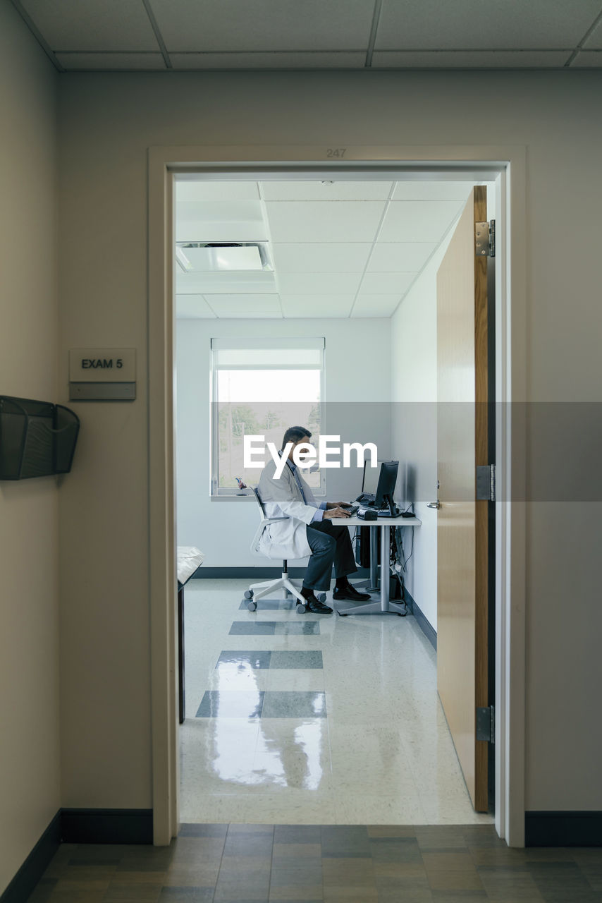 Side view of doctor using desktop computer while working in hospital seen through doorway